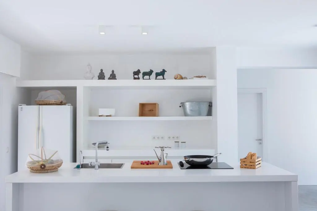 Bright white kitchen with open shelving displaying decorative items. A white counter features a sink, cooktop, and cooking utensils.