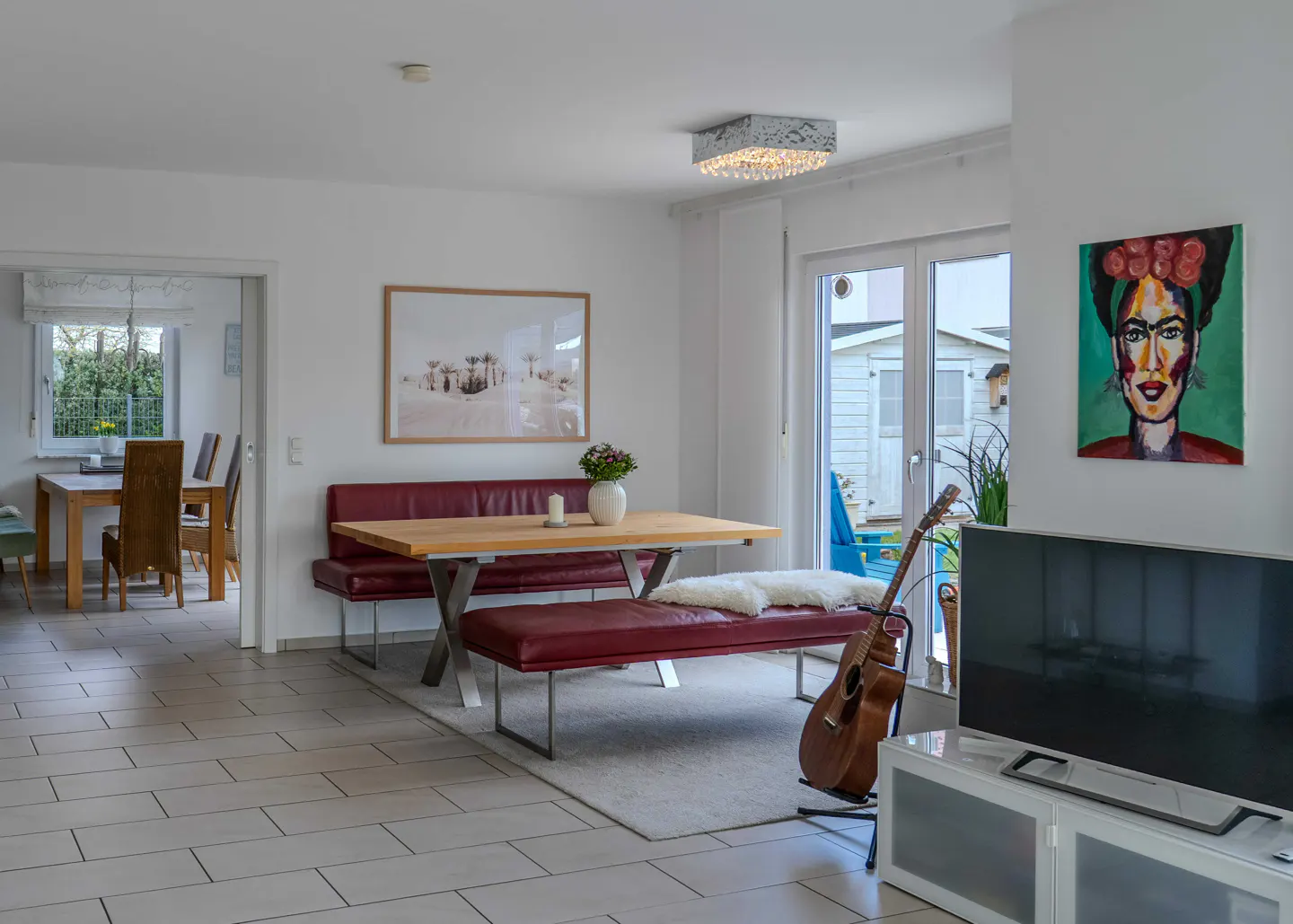 Bright, modern dining room with a wooden table, red benches, and a Frida Kahlo portrait on the wall. A guitar leans against the bench.