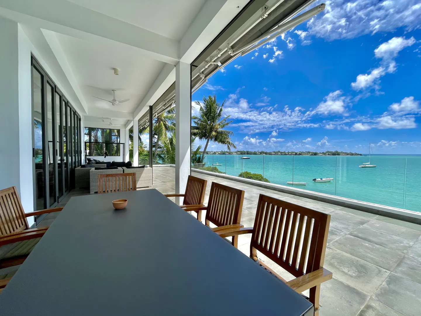 Outdoor patio with a long table and chairs overlooking a turquoise ocean with boats under a blue sky.