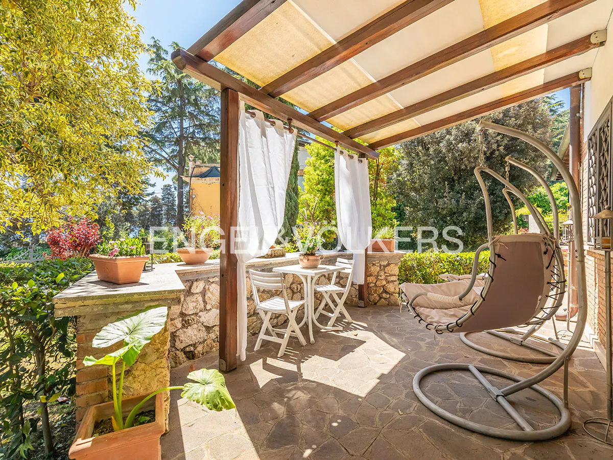 Outdoor patio with a swing chair, table, and chairs under a pergola with white curtains. Lush greenery surrounds the space.