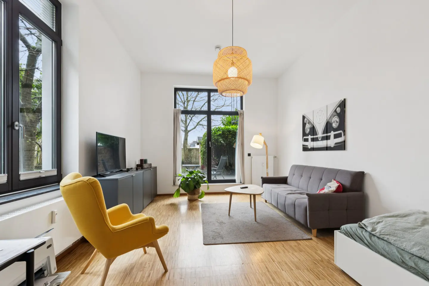 Bright living room with wood floors, white walls, and a gray sofa. A yellow chair sits near a window. A woven pendant light hangs from the ceiling.