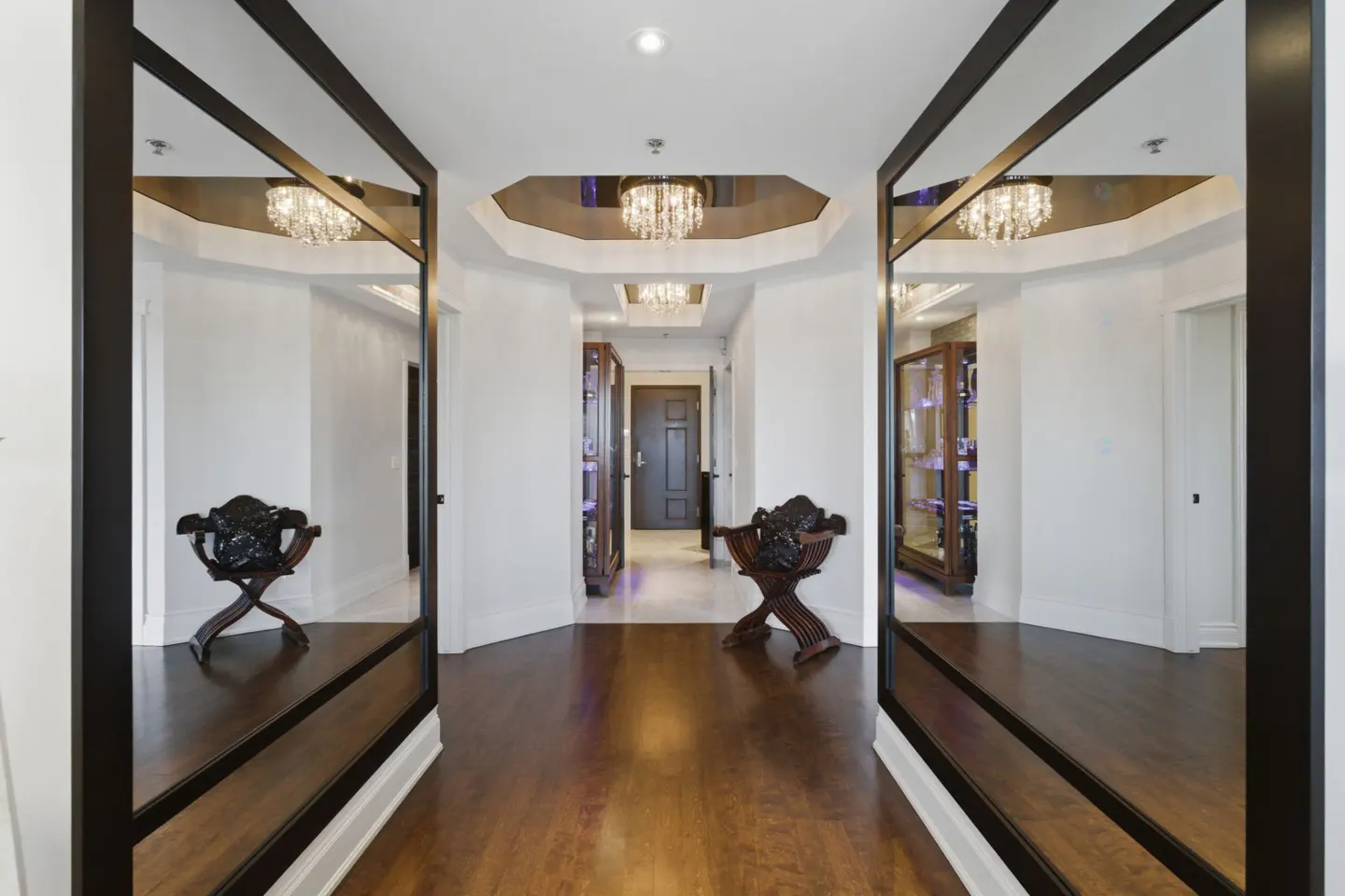 Hallway with dark wood floors, white walls, and black-framed mirrors. A dark wood chair sits between the mirrors. Chandeliers hang from the ceiling.