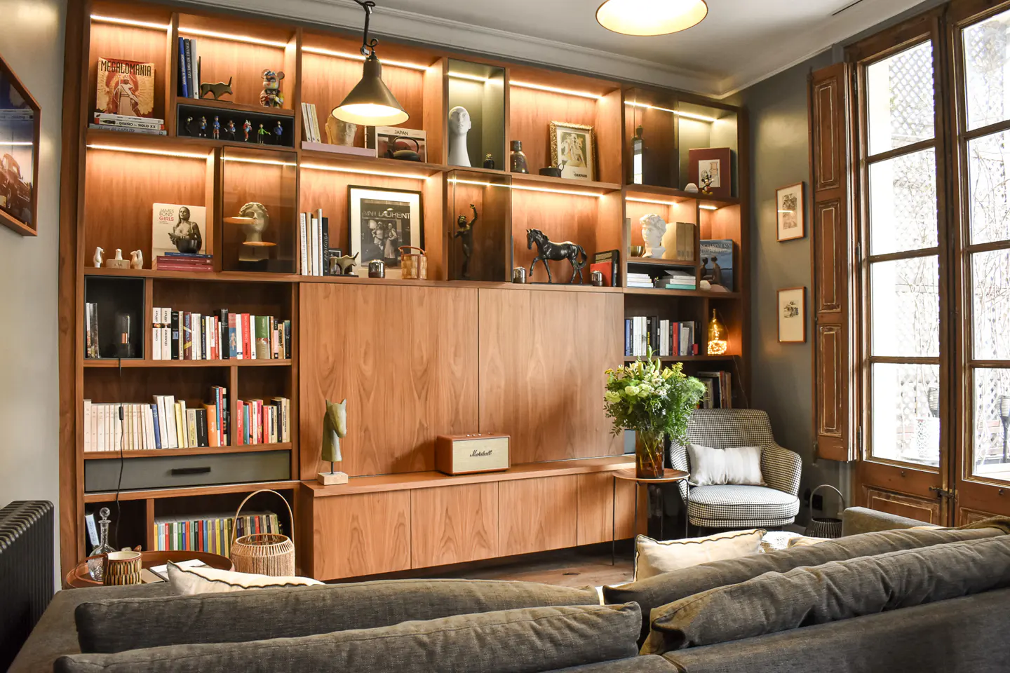 Living room with a large wooden bookcase filled with books and decorative objects, illuminated by overhead lighting. A gray sofa is in the foreground.