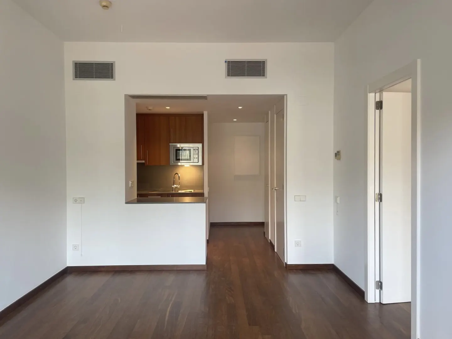 An interior view of a modern apartment with white walls, dark wood floors, and an open kitchen with brown cabinets.
