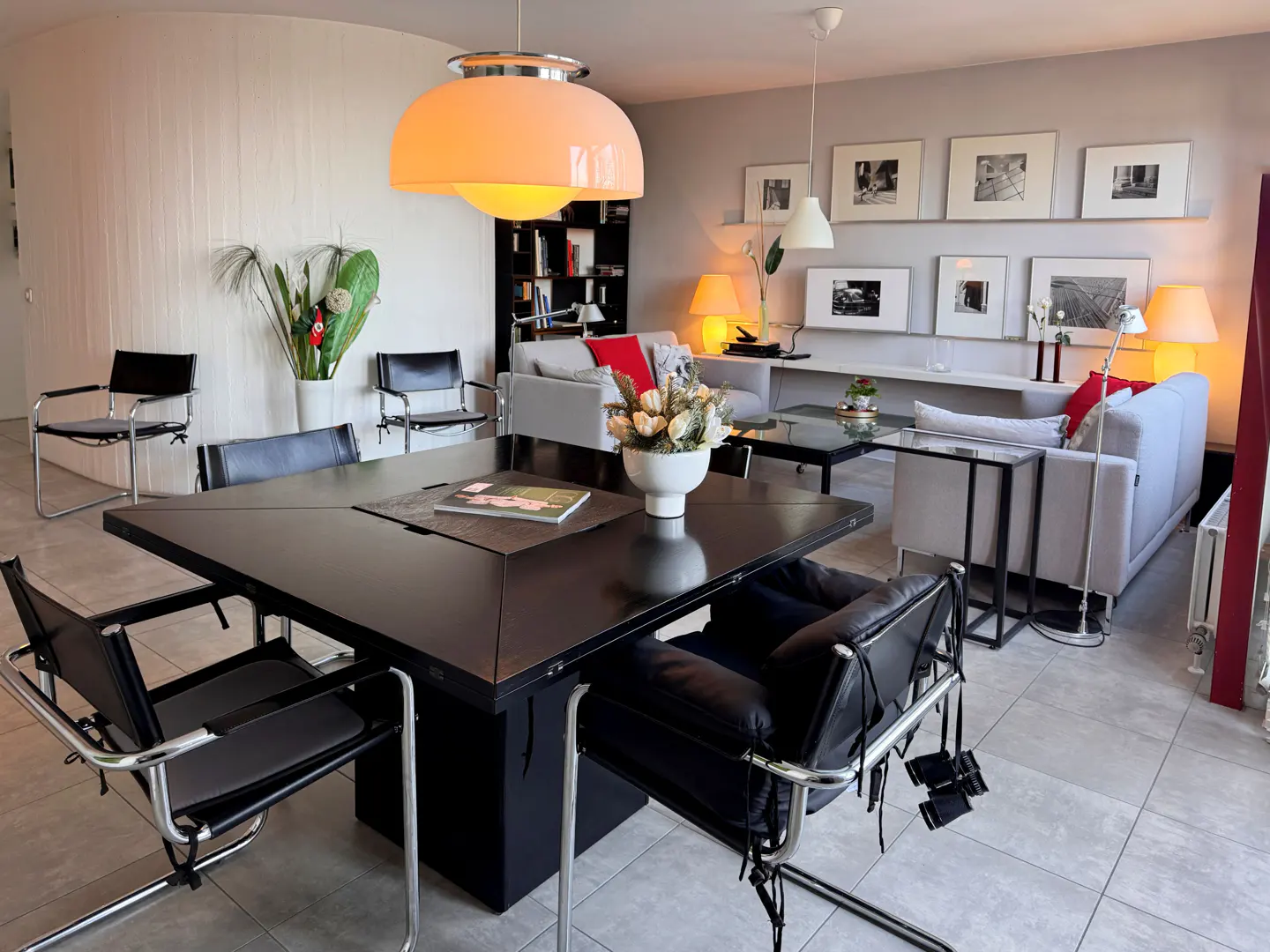 A modern living room with a black square table, chrome chairs, and a gray sofa with red pillows. A large orange pendant light hangs above.