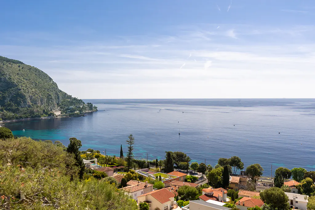 Scenic view of the French Riviera featuring red-roofed houses, lush greenery, and the blue Mediterranean Sea under a clear sky.