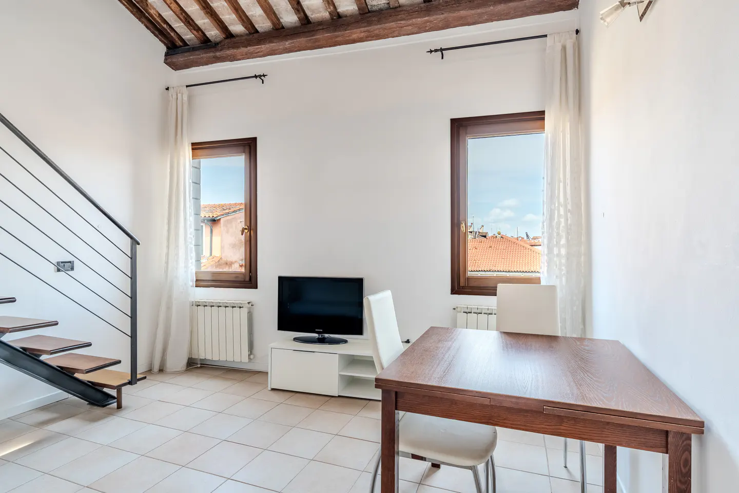Bright living room with a wooden table, white chairs, and a TV. Windows show rooftops. Stairs lead up. Beamed ceiling.