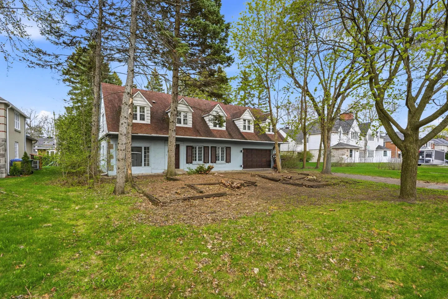 A light blue two-story house with a brown roof and dormer windows, surrounded by trees and a green lawn.