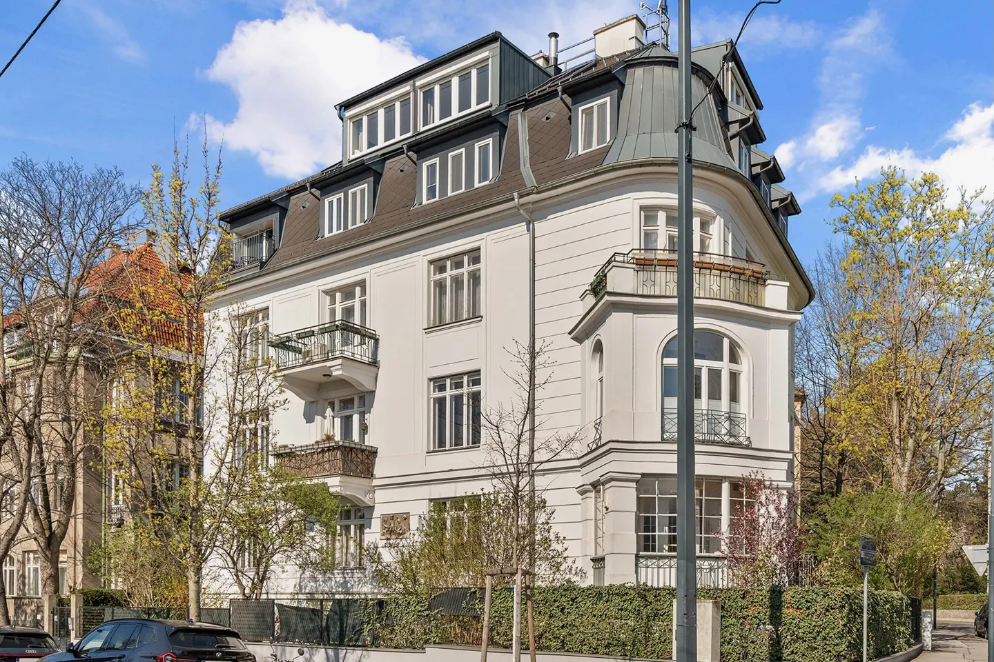 Exterior view of a three-story white building with a gray roof and balconies, surrounded by trees and a hedge.