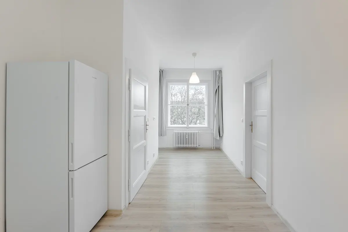 Bright, white hallway with light wood floors. A white refrigerator is on the left, and a window is at the end of the hall.