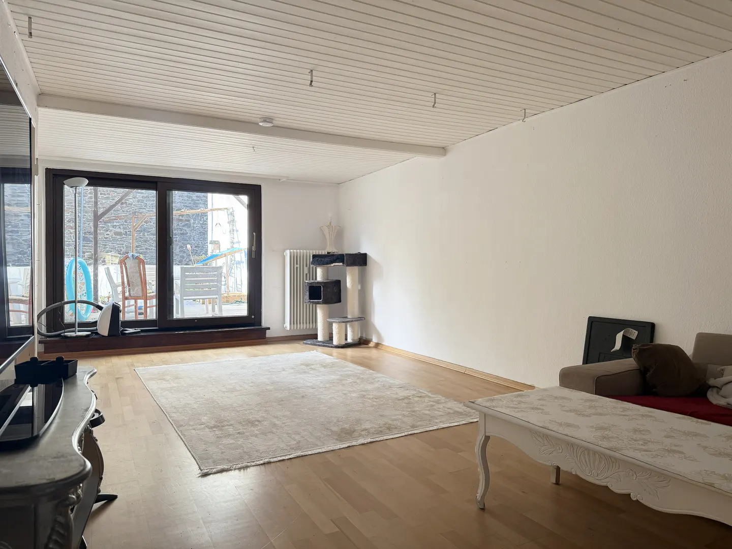 Bright living room with wood floors, white walls, and a large window. A beige rug sits in the center, with a cat tree and white coffee table nearby.