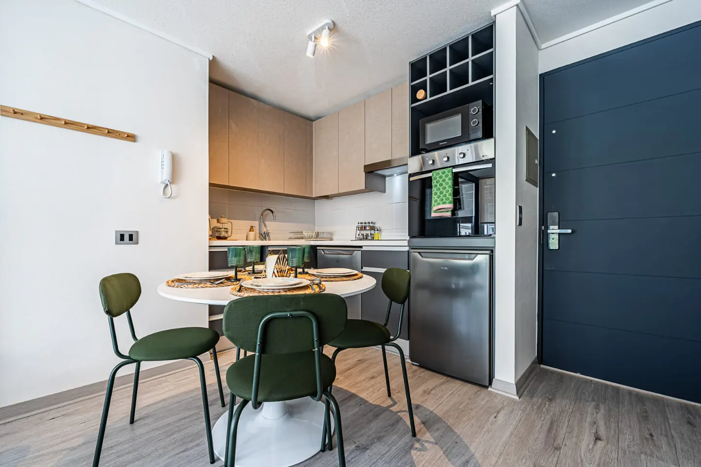 Modern kitchen with light wood cabinets, stainless steel appliances, and a round dining table with three green chairs.