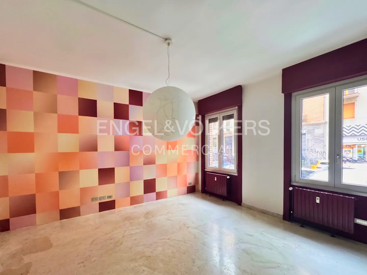 Empty room with marble floors, a colorful square-patterned wall, and two windows with burgundy trim and radiators. A white globe light hangs from the ceiling.