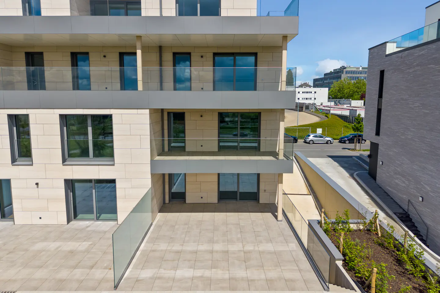 Exterior view of a modern, multi-story apartment building with large windows and balconies. A tiled patio is visible on the ground floor.