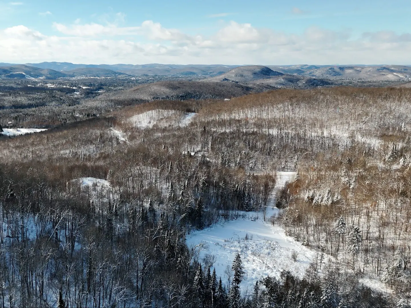 Aerial view of snow-covered hills and forests under a blue sky with scattered clouds.