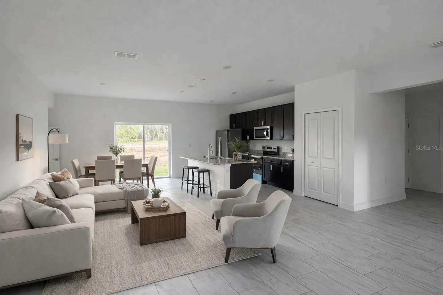 Open-concept living space with white walls, gray tile floors, and dark kitchen cabinets. Beige sectional sofa and chairs.