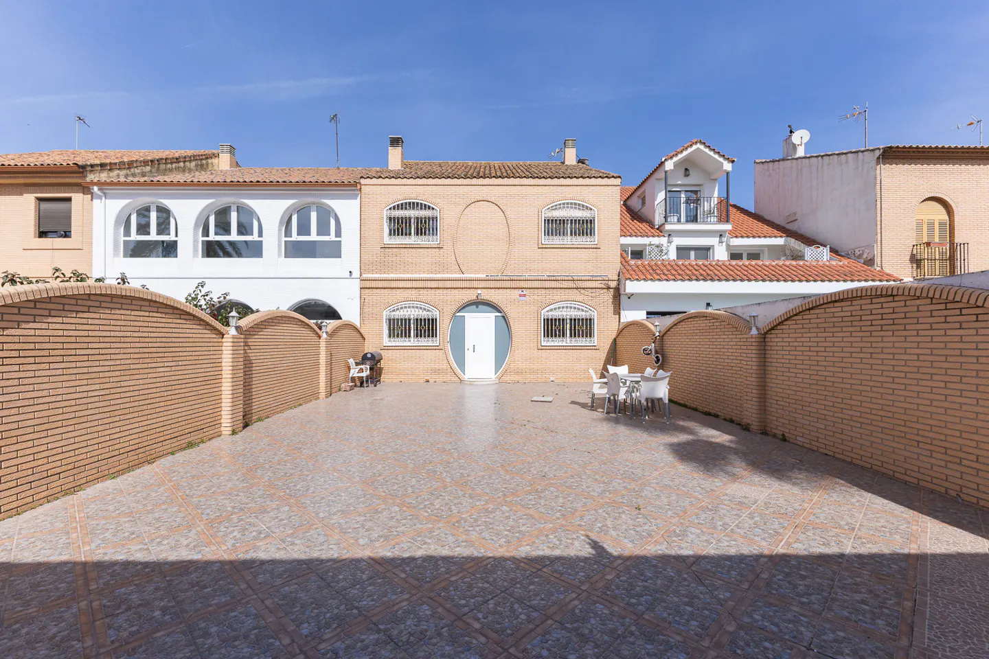 Exterior view of a tan brick house with a white door and barred windows, surrounded by a brick wall and tiled patio.