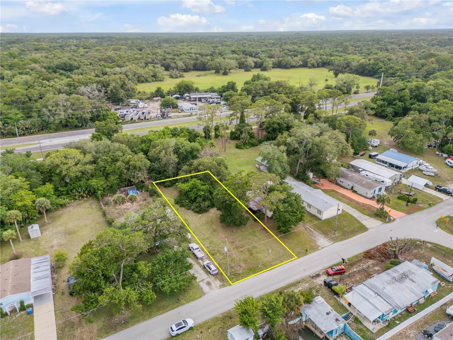 Aerial view of a vacant lot outlined in yellow, surrounded by trees, homes, and a road. A green field and forest are in the background.