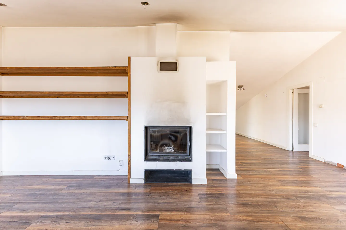 Bright living room with dark wood floors, white walls, and a fireplace. Wooden shelves are on the left, and white shelves are on the right.
