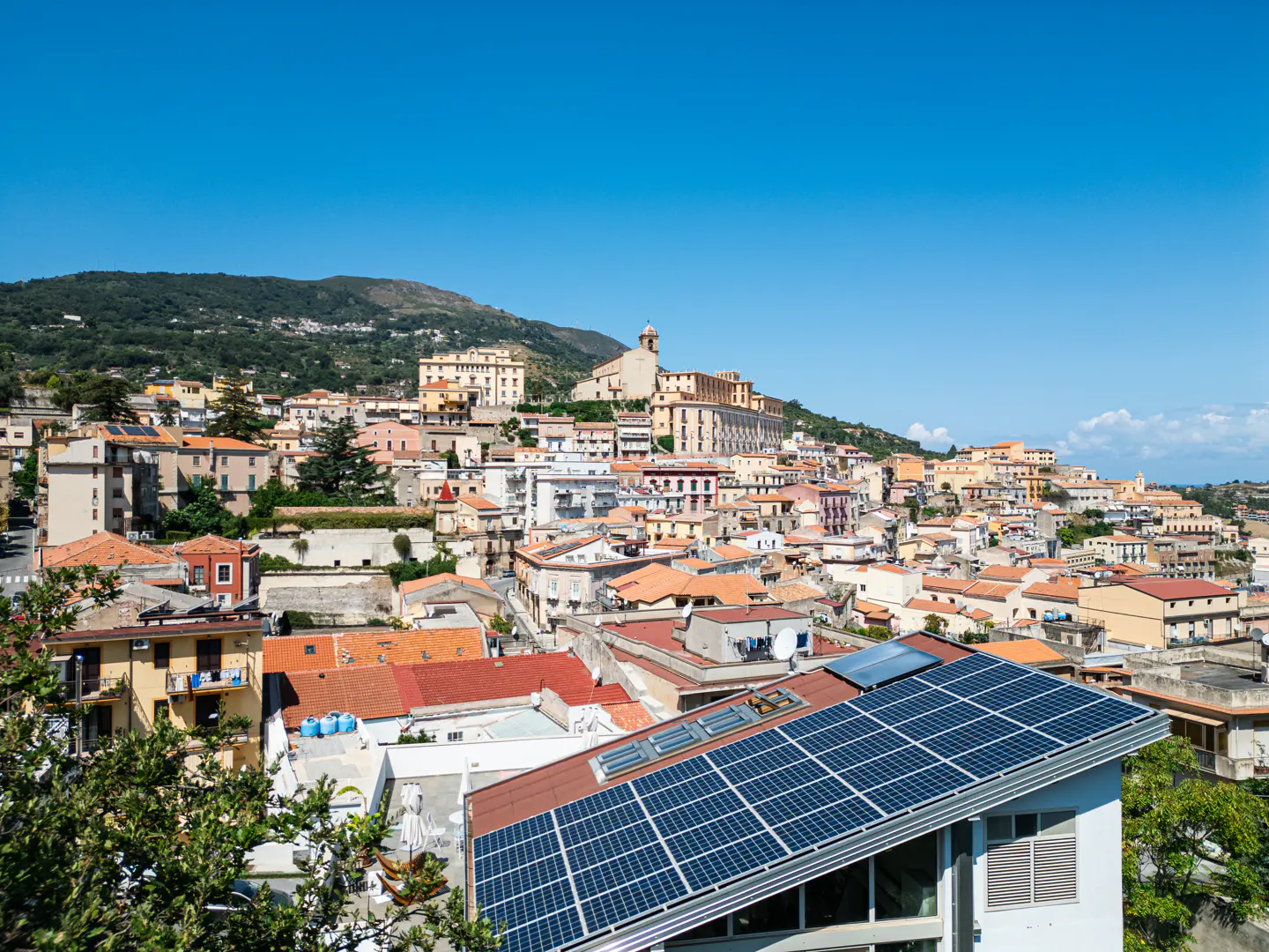 View of a hillside town with red-tiled roofs, a church, and a house with solar panels under a clear blue sky.