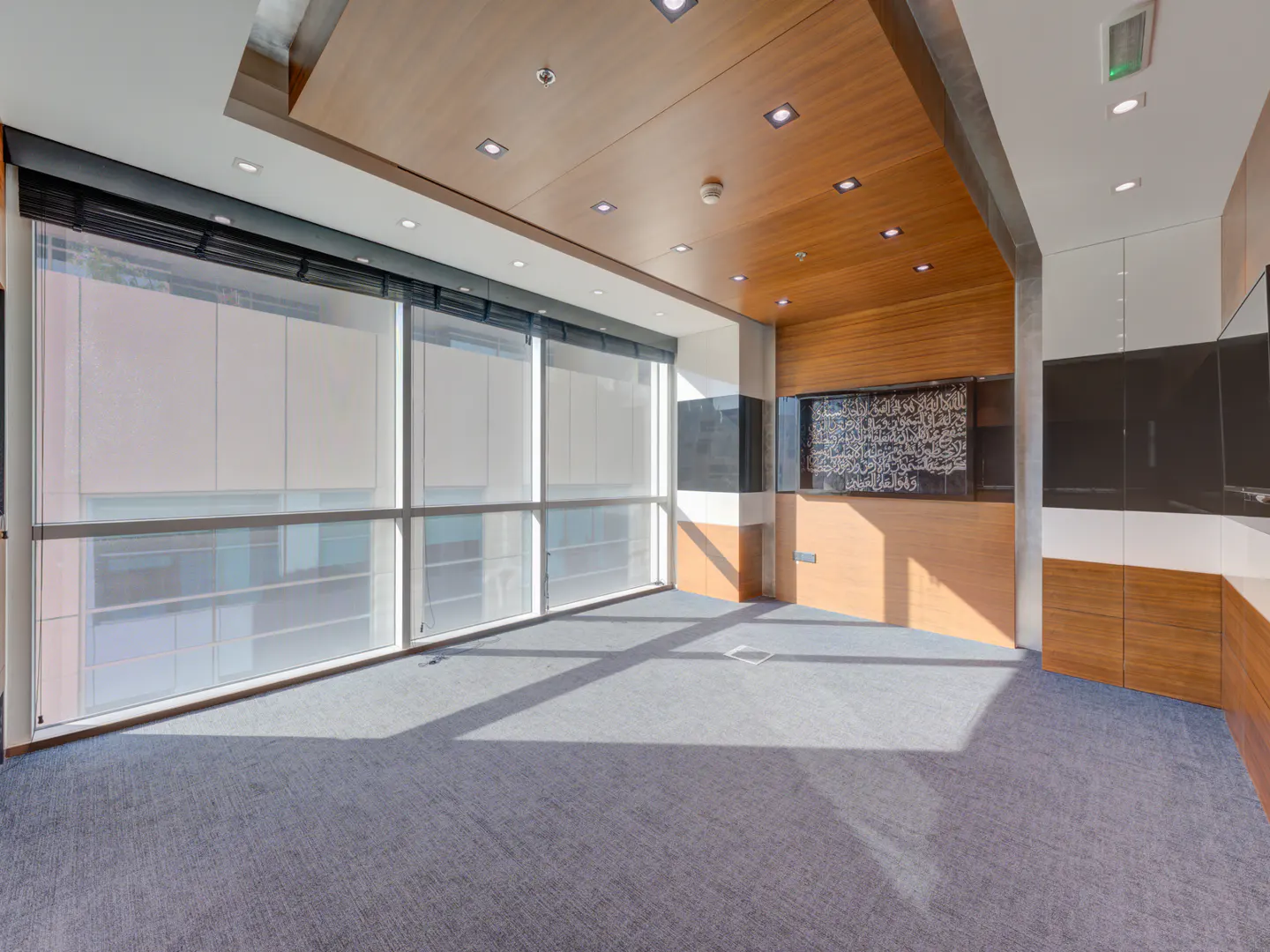 Empty office space with gray carpet, large windows with blinds, and wood-paneled walls and ceiling. Recessed lighting illuminates the room.