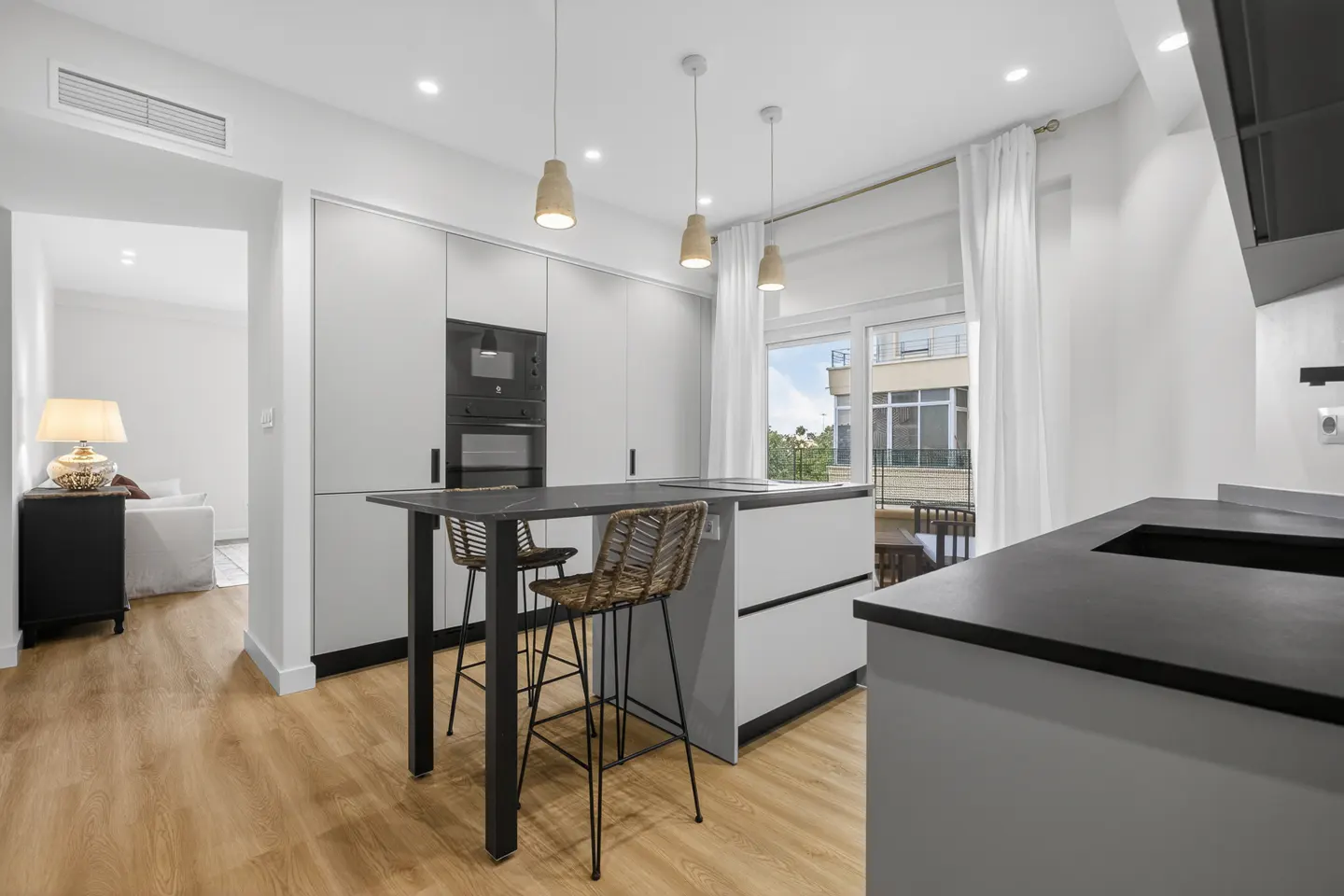 A modern kitchen with white cabinets, black countertops, and a kitchen island with two woven bar stools. Three pendant lights hang above the island.