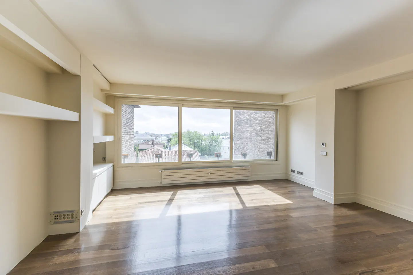 Bright, empty room with hardwood floors, white walls, and a large window overlooking a cityscape. Built-in shelving on the left.