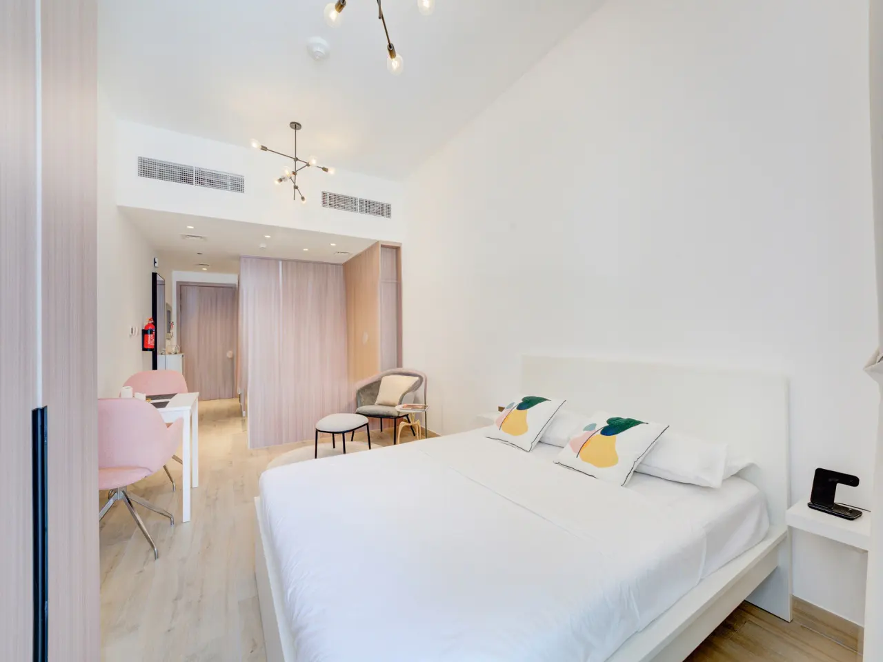 Bright studio apartment with white walls, a bed with patterned pillows, and a small seating area with pink chairs.