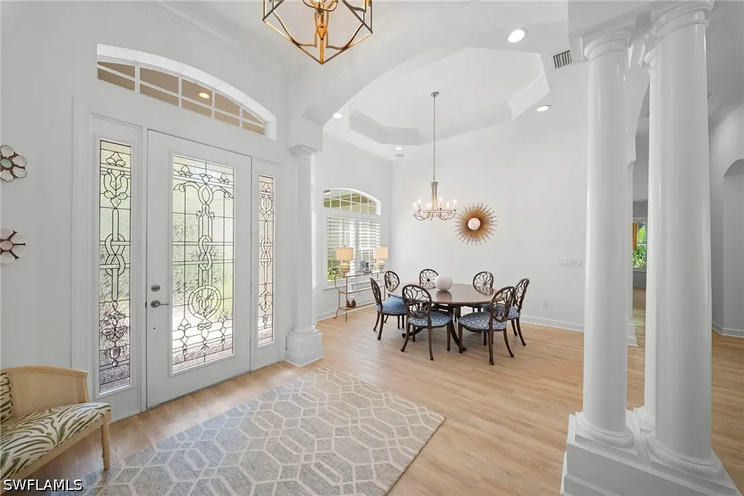 Bright foyer with white walls, light wood floors, and a geometric rug. A dining table with six chairs sits under a chandelier. Stained glass front door.