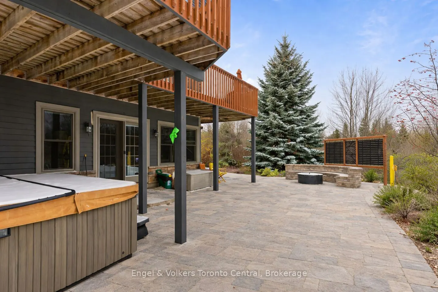 Backyard patio with a hot tub, stone pavers, fire pit, and a large evergreen tree. The house has a gray exterior and a wooden deck.