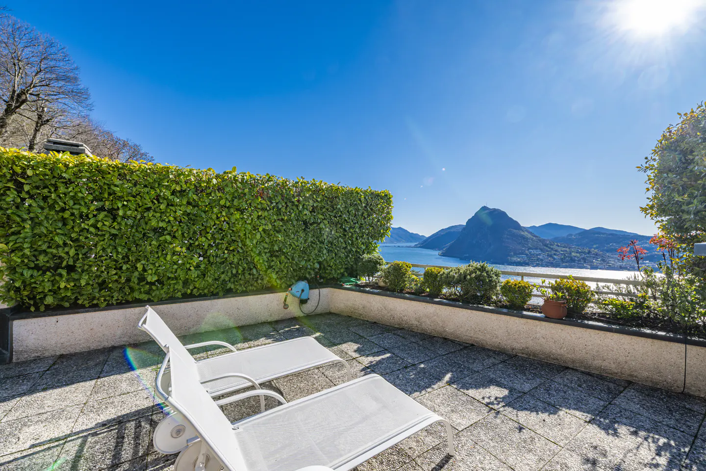 Sunny patio with two white lounge chairs, green hedge, and a view of a lake and mountains under a clear blue sky.