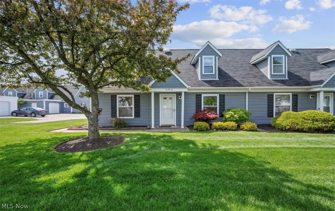 Exterior view of a blue, two-story townhouse with a white door, black shutters, and a green lawn. A large tree is on the left.