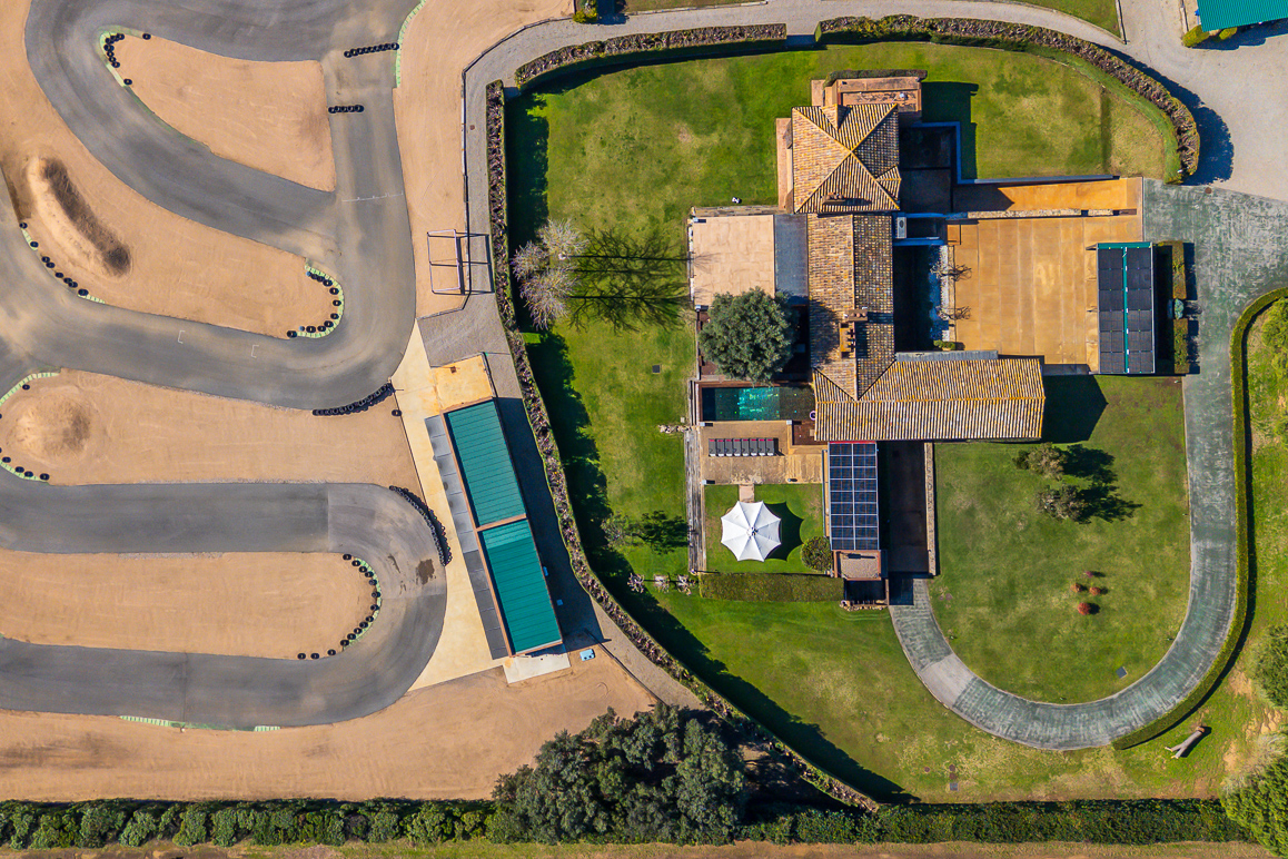 Aerial view of a house with a tiled roof, pool, and white umbrella, situated next to a winding dirt go-kart track. [AI-generated text]