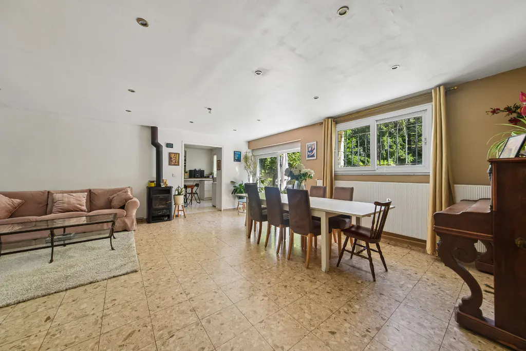 A spacious living and dining area with a brown sofa, wood-burning stove, white table with brown chairs, and a dark wood piano.