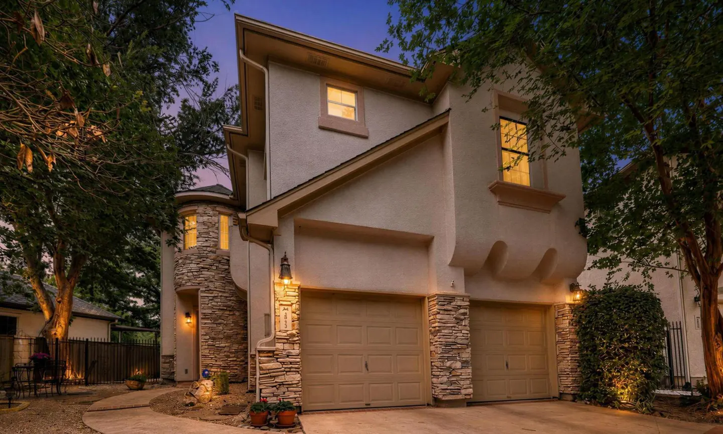 Two-story stucco home at dusk with stone accents, a two-car garage, and warm exterior lighting surrounded by trees.