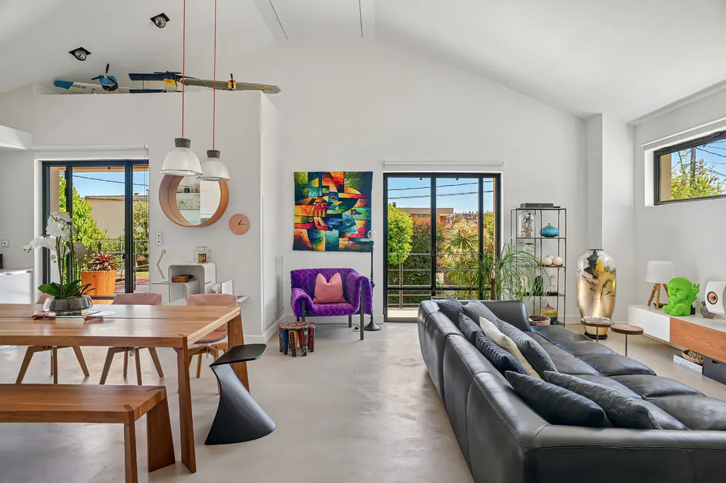 Bright, modern living room with a wood table, black sofa, purple chair, and colorful art. Large windows offer views of greenery.