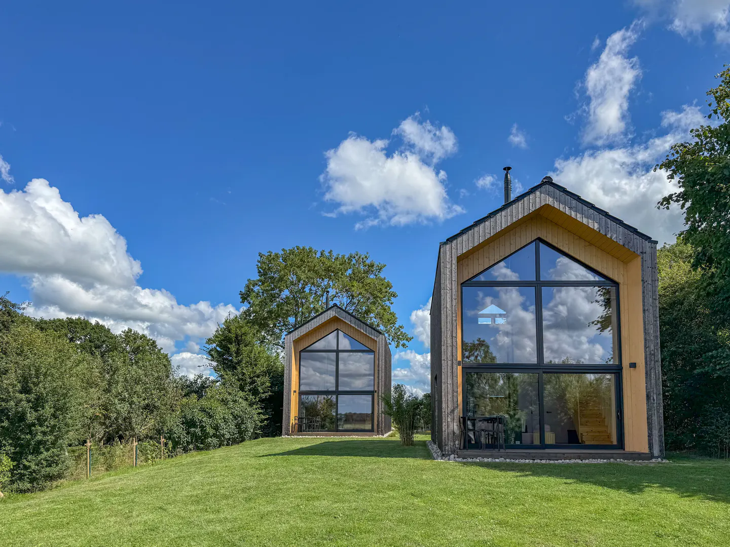 Two modern, wood-clad cabins with large windows sit on a green lawn under a blue sky with scattered clouds. Trees surround the property.