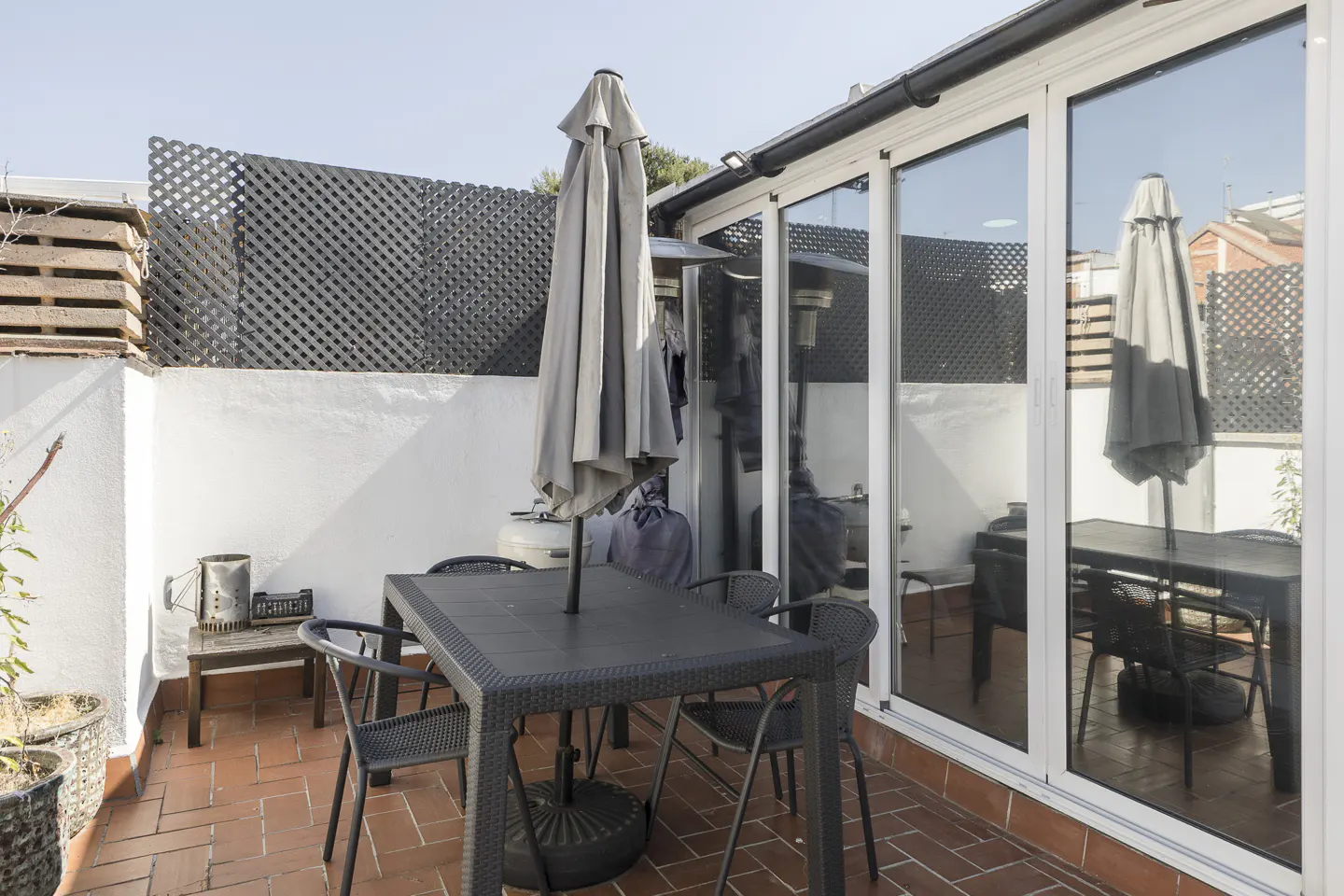 Outdoor patio with a black table, chairs, and closed gray umbrella on a brick floor. White walls and sliding glass doors are visible.