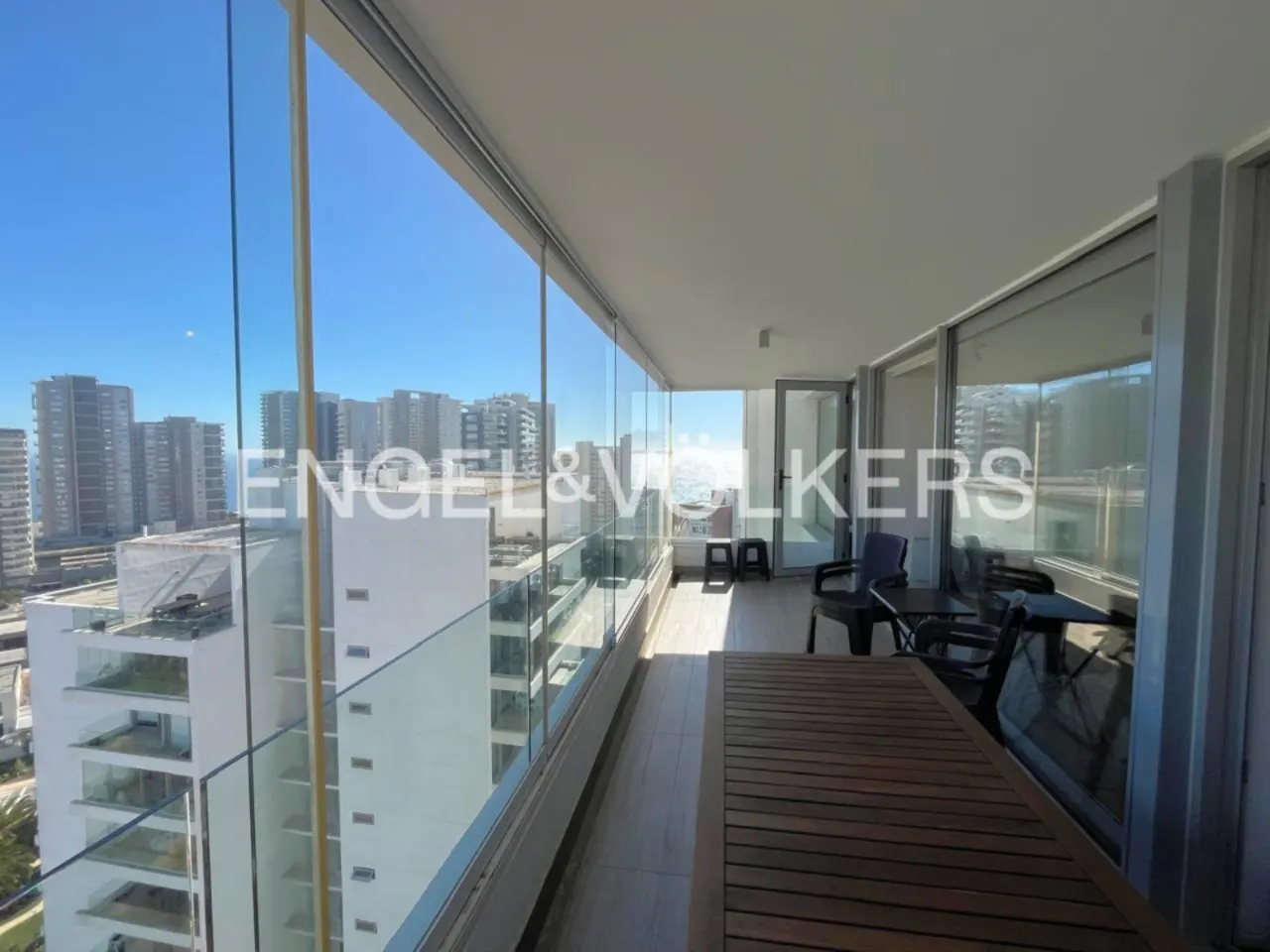 A bright balcony with a wooden table and chairs overlooks a city skyline and ocean through glass railings. The Engel & Volkers logo is visible.