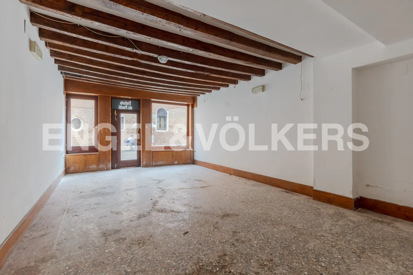 Empty room with white walls, a concrete floor, and exposed wooden beams on the ceiling. A storefront window is visible.