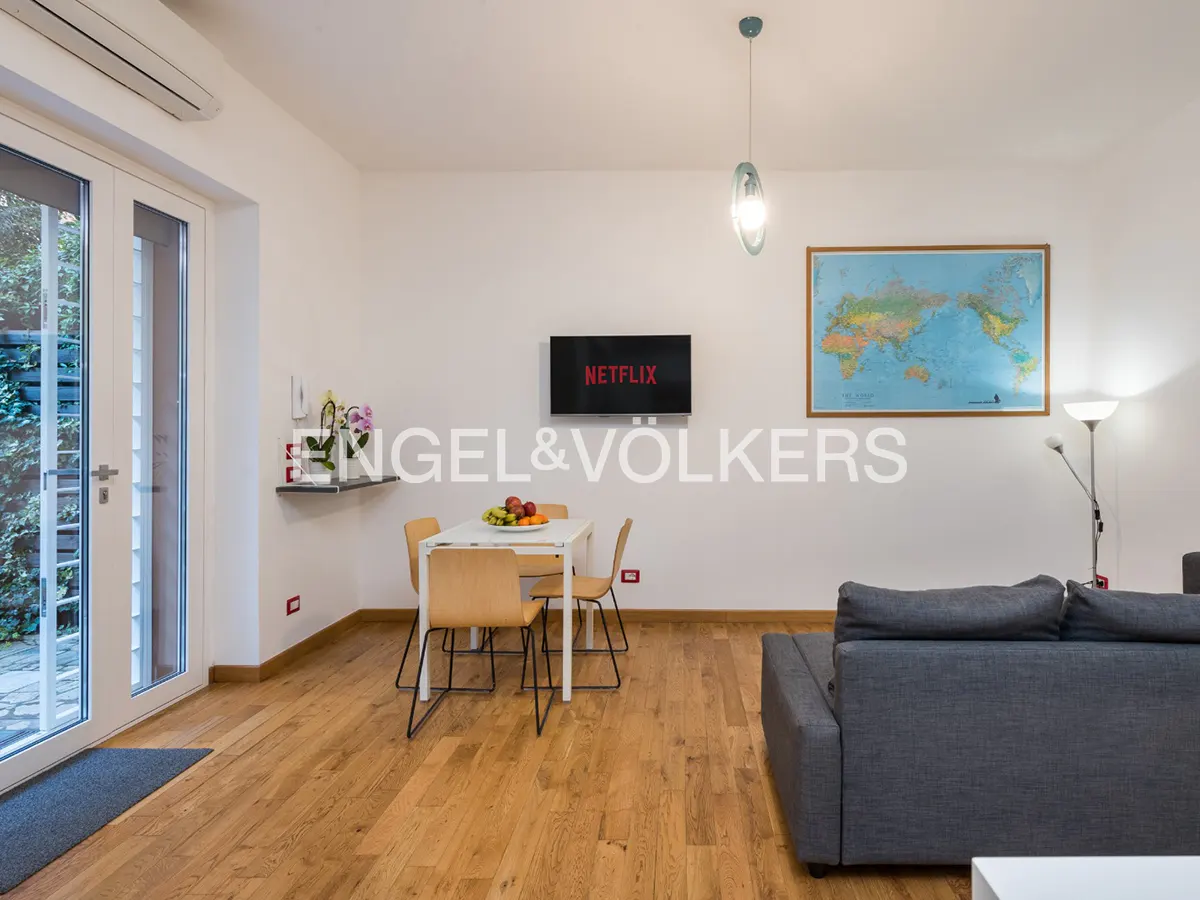 Bright living room with wood floors, white walls, and a gray sofa. A dining table sits near a TV with the Netflix logo. A world map hangs on the wall.