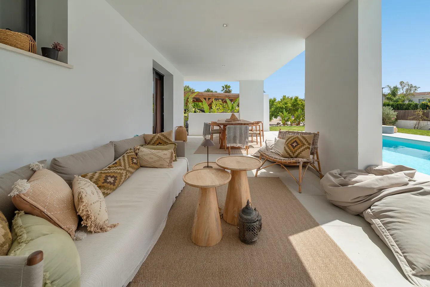Covered patio with a long sofa, wooden tables, and a wicker chair. A dining table is visible in the background, with a pool to the right.