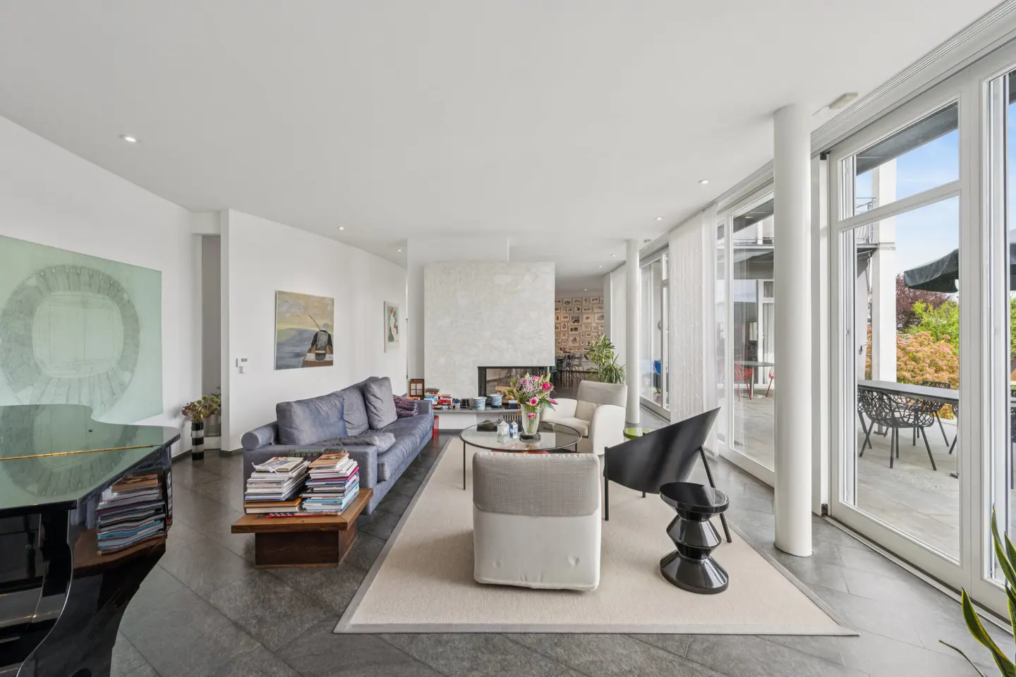 Bright living room with a blue sofa, fireplace, piano, and a wall of windows leading to a balcony.