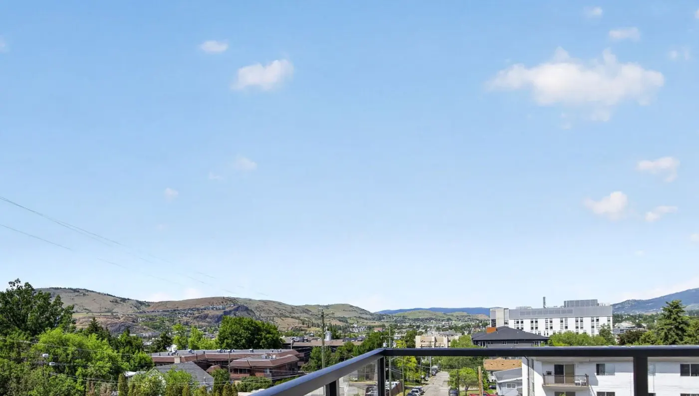 View from a balcony overlooking a suburban town with green trees, buildings, and distant mountains under a blue sky.