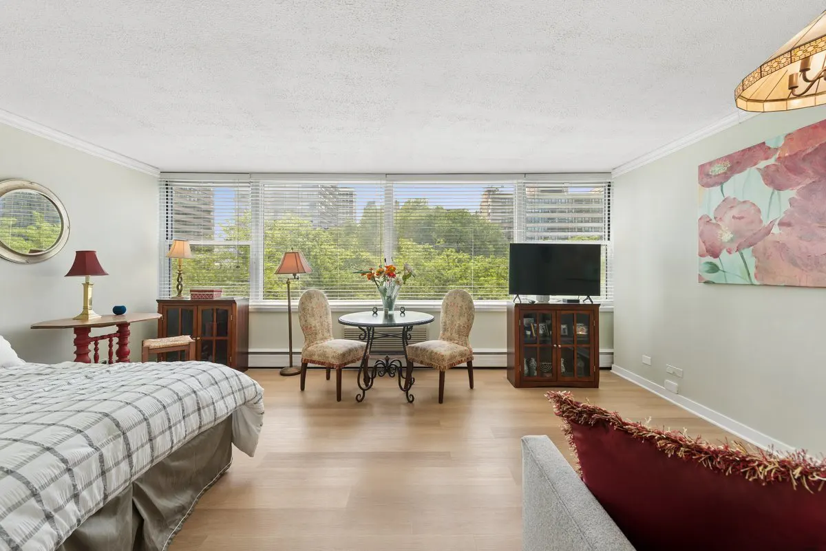 Bright studio apartment with a bed, table and chairs, TV, and a large window with a view of trees and buildings.