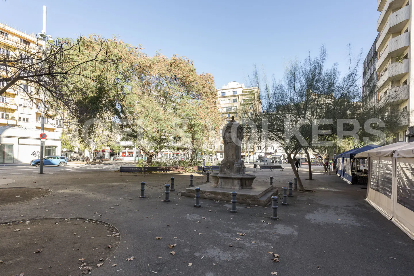 City park with a stone fountain in the center, surrounded by trees and buildings under a clear blue sky. Market stalls line the right side.