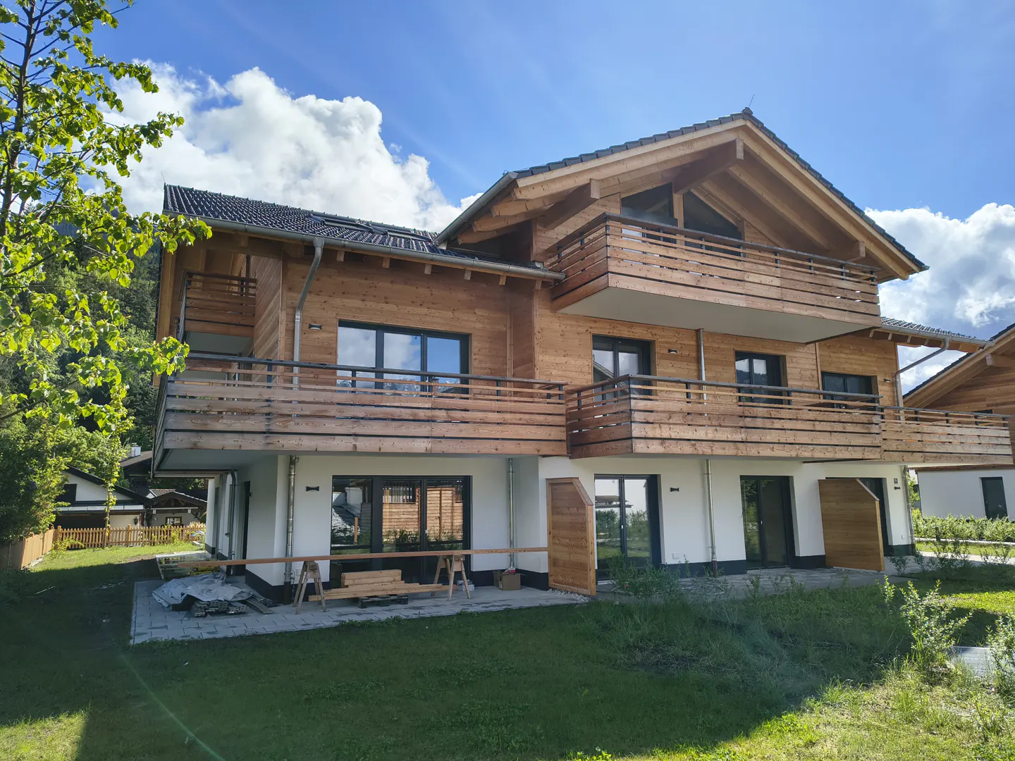 Two-story wood house with balconies, white base, dark roof, and green lawn under a blue sky with clouds.