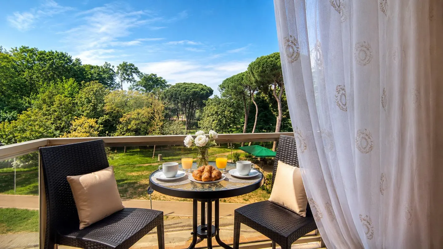 A balcony table with coffee, croissants, and flowers overlooking a lush green park on a sunny day.