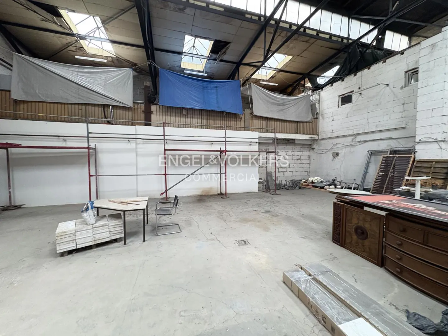 Large, empty commercial space with scaffolding, a table, and furniture. Skylights and tarps are visible in the ceiling. "Engel & Volkers Commercial" is on the wall.