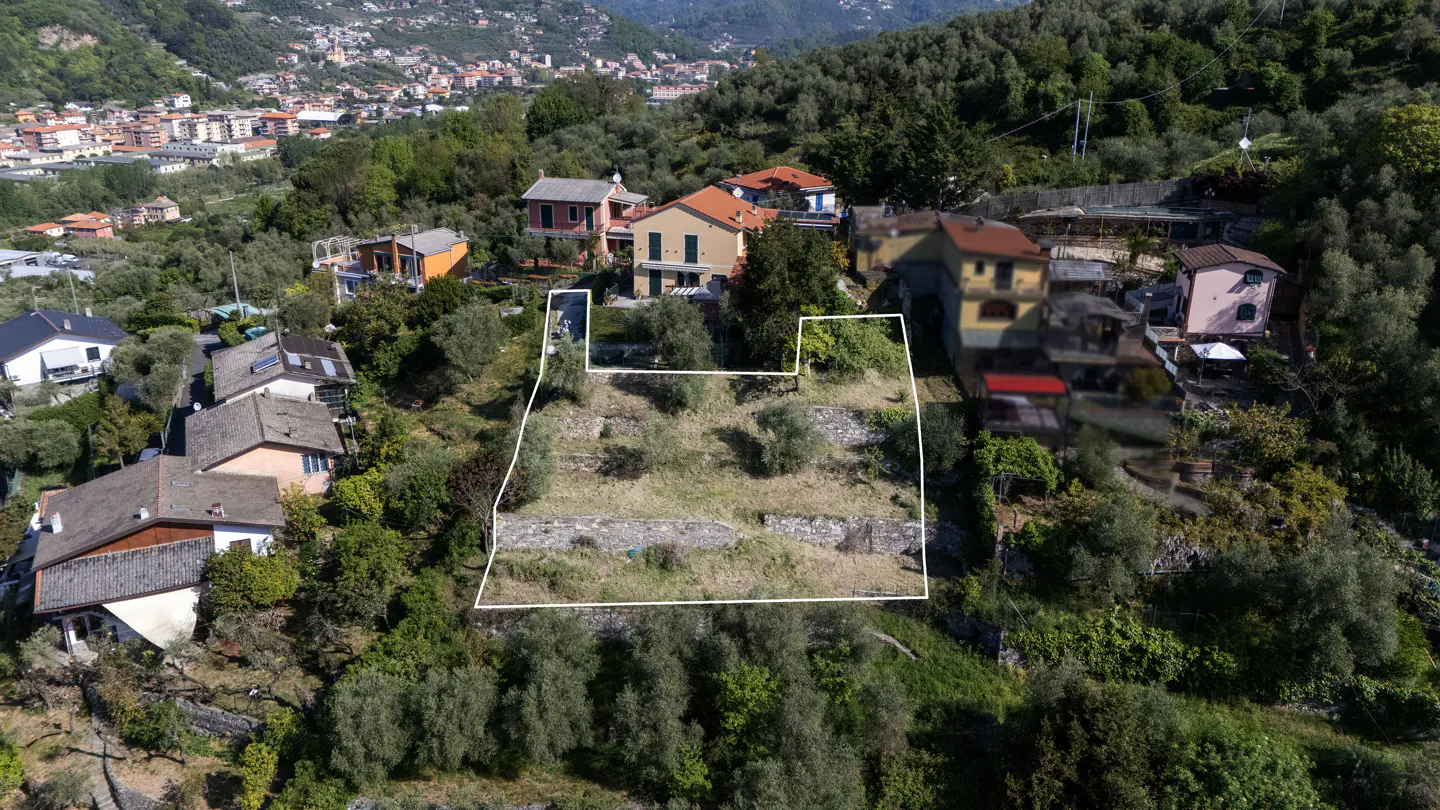 Aerial view of a vacant lot outlined in white, on a hillside with trees and houses in the background.